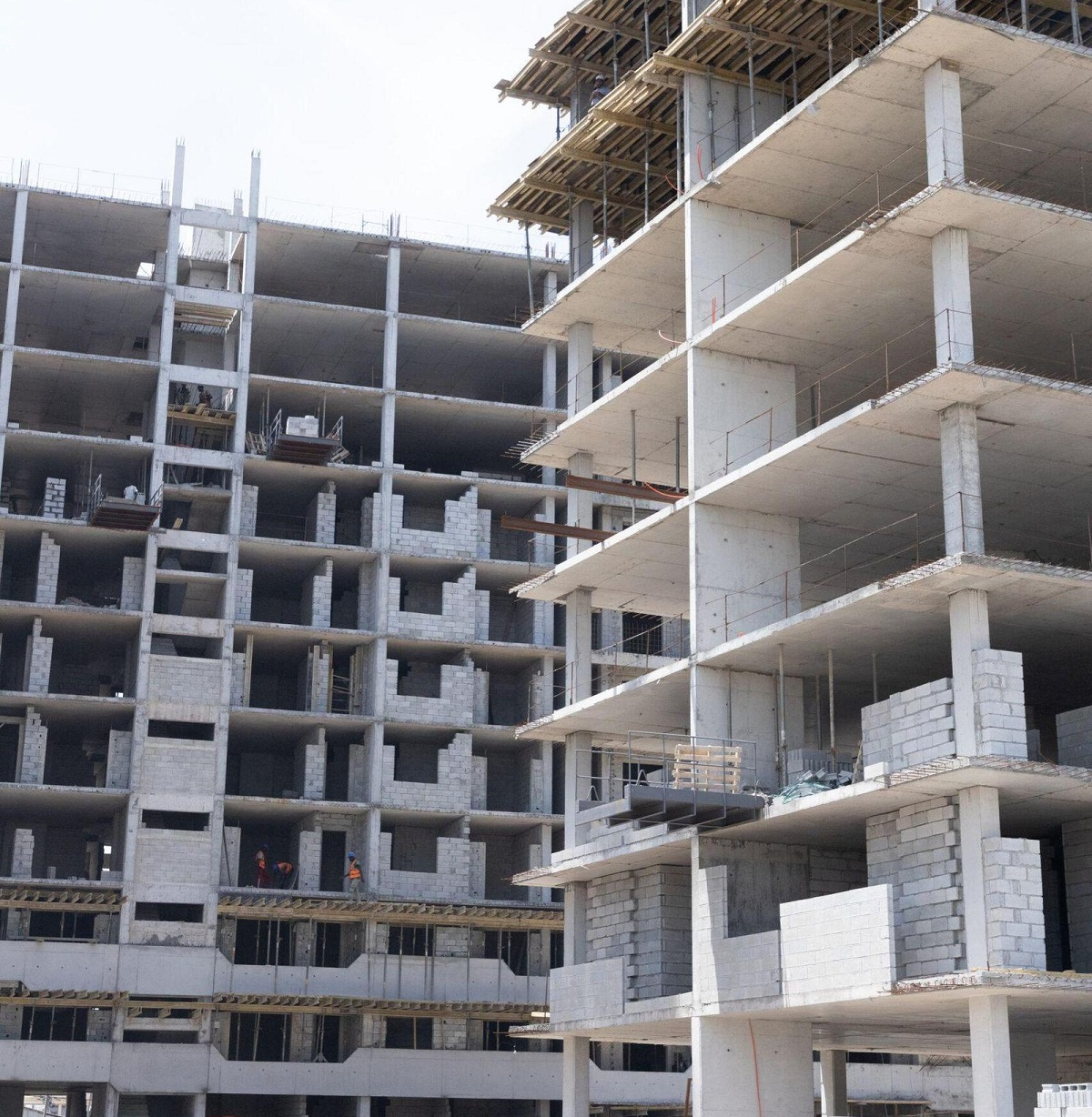 A high-angle photograph showing a multi-story concrete building under active construction, highlighting the upper floors. The design utilizes thin post tension slabs which eliminate traditional deep support beams and maximize clear ceiling height, allowing for more floors. Concrete block infill and construction equipment are visible at the lower levels.